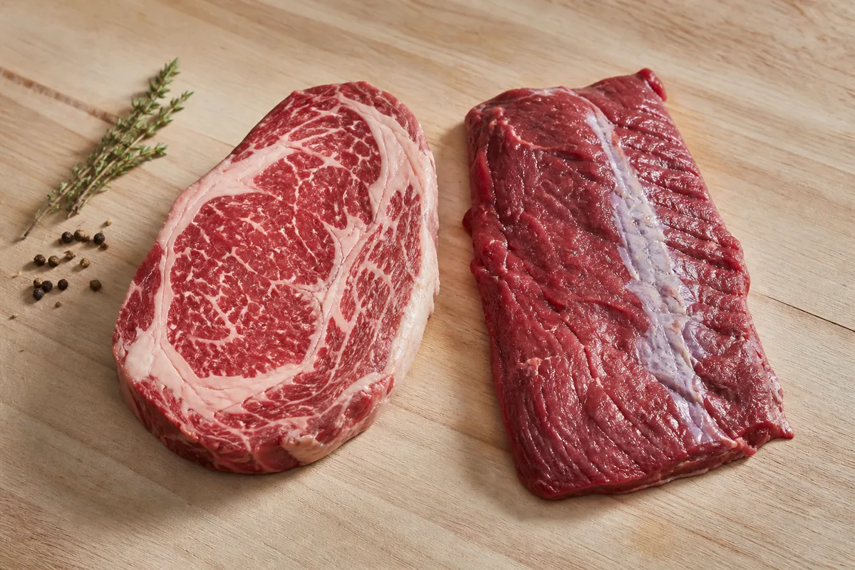Deux morceaux de boeuf crus posés sur un billot de boucher, une pièce persillée à griller et une pièce à braiser plus fibreuse